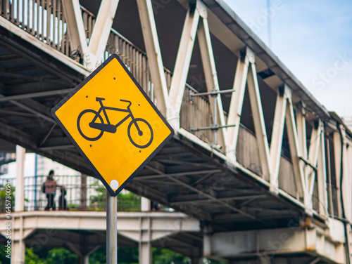 Close-up of a yellow bicycle road sign in front of a pedestrian bridge, highlighting urban infrastructure and cycling safety in the city.