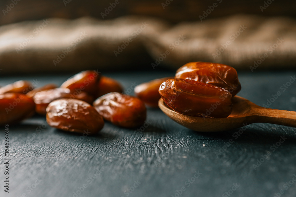 Obraz premium Scattered dried dates on a dark background with a wooden spoon. A healthy, naturally sweet snack full of energy and nutrients, often enjoyed during Ramadan and in Middle Eastern cuisine