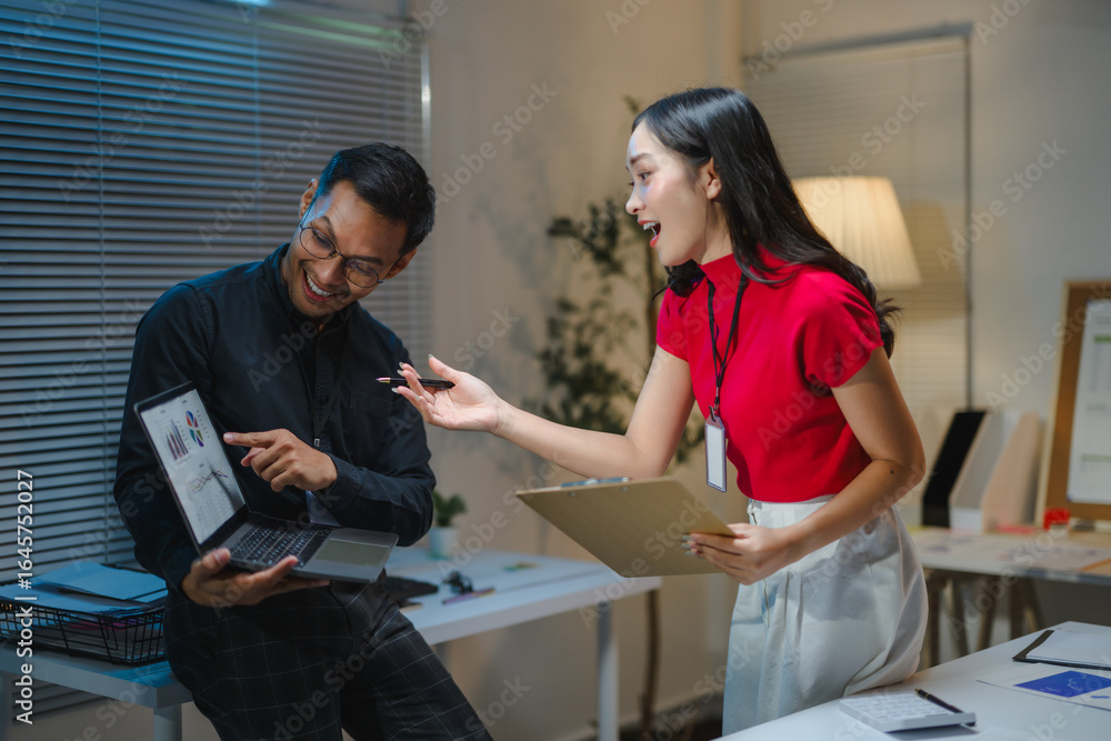 Fototapeta premium Asian businesspeople discussing positive financial charts on laptop in office at night