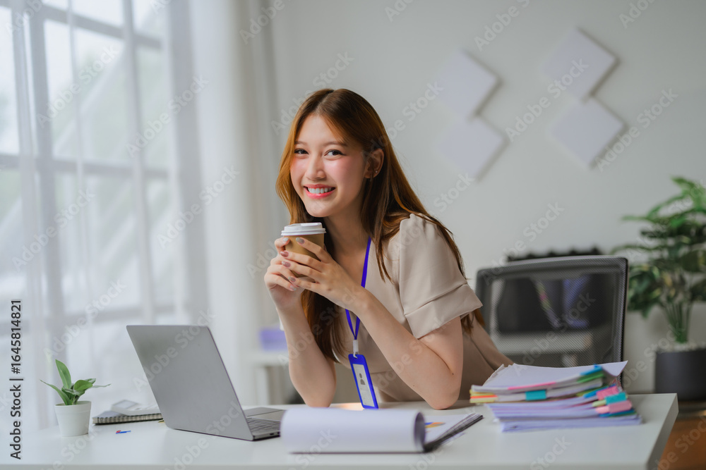 Obraz premium Young businesswoman smiling and holding coffee cup while working on laptop in modern office