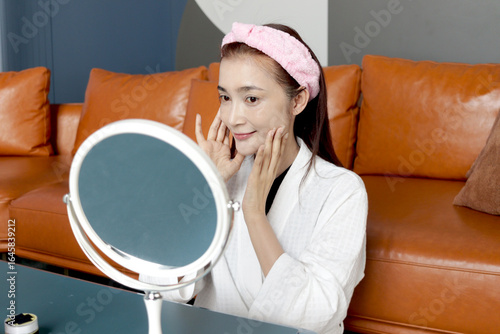 Happy middle aged Asian woman in bathrobe look in mirror, apply cosmetic on face. Beautiful adult female treatment her face with skincare hydrating at home. Beauty healthcare treatment and cosmetics.