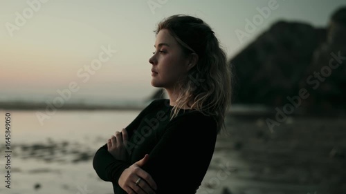 A thoughtful woman stands with arms crossed, gazing into the distance at sunset on a beach. The serene atmosphere evokes contemplation and introspection.