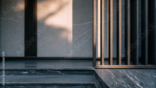 A close-up of modern architectural details with a textured grey wall and dark vertical slats. Sunlight highlights the minimalist design and dark stone steps.

