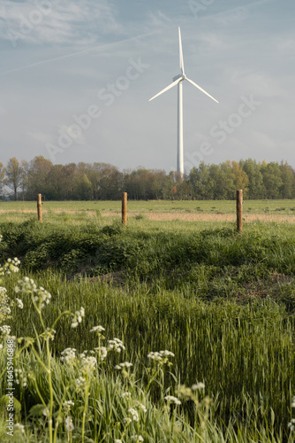 Vertical spring landscape in Flevoland, Netherlands, with wildflowers, green grass, trees on the horizon, and a wind turbine in the distance under a clear blue sky.