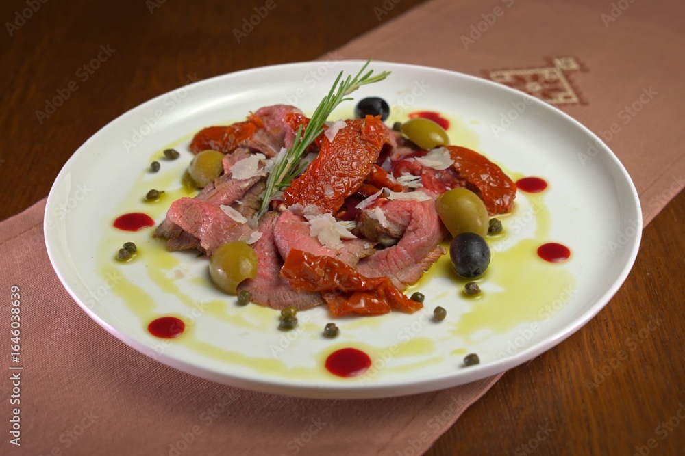 Beef carpaccio with sun-dried tomatoes, olives, capers, parmesan shavings, and rosemary, drizzled with olive oil and berry reduction on a white plate. Beef carpaccio with sun-dried tomatoes, olives, capers, parmesan shavings, and rosemary, drizzled with olive oil and berry reduction on a white plate.