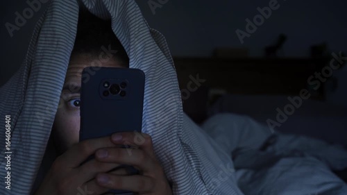 Smartphone and social network addiction concept. Young male using smartphone in bed with blanket over head