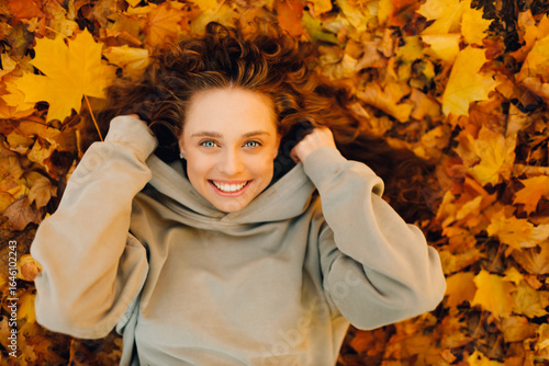 Smiling young woman lies on the autumn foliage in the park with the yellow leaves