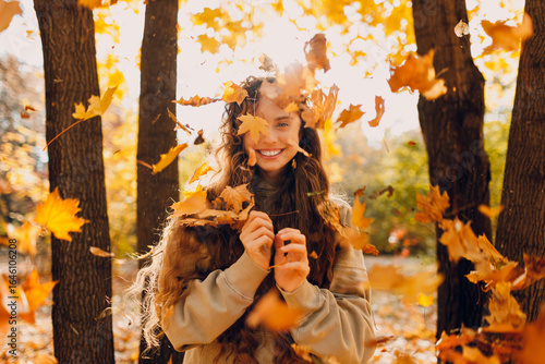 Smiling positive young woman throws up the yellow autumn leaves in the forest at sunset