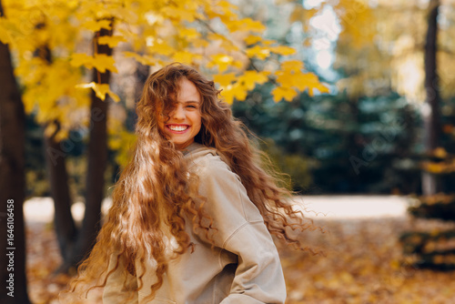 Smiling young woman with curly hairstyle enjoys the autumn weather in the forest with the yellow leaves at sunset