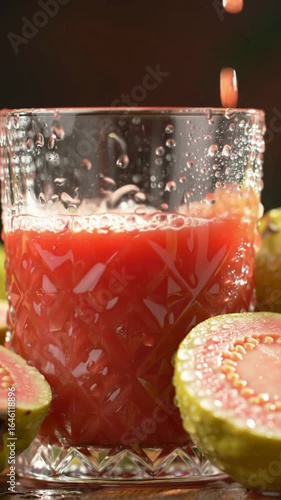 Fresh pink guava juice being poured into a crystal glass, tropical fruit halves in foreground with water droplets