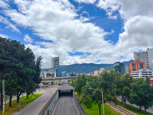 Vibrant panoramic view of Bogotá, Colombia, showcasing Calle 100 with modern buildings, green trees, and mountains in the background under a partly cloudy sky