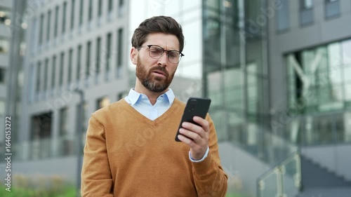 Shocked worried businessman reading bad news on mobile phone while standing on the street near an office building. Frustrated stressed entrepreneur worker has a bad mood, looking at negative message