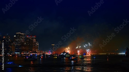 Water vehicles are standing still on the waterscape of the river in New York. Firework show is taking place over the water.
