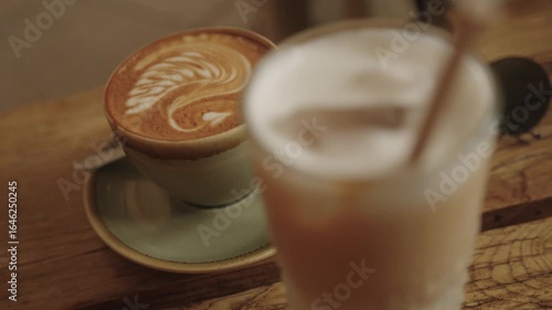 Cappuccino with latte art and blurred iced latte in the foreground on a rustic wooden café table