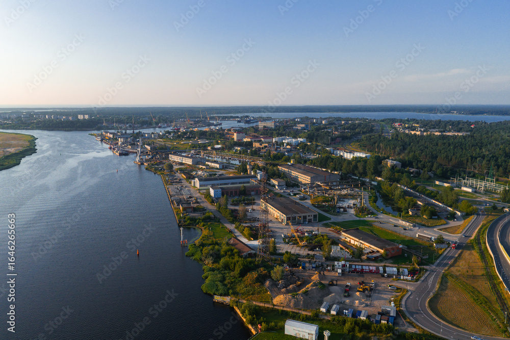 Naklejka premium Aerial view of a shipyard and industrial zone by the Daugava River in Riga, Latvia, with docks, cranes, and urban structures under a clear blue sky.