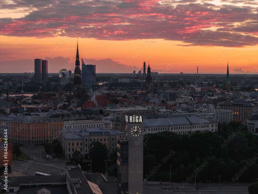 Fototapeta premium Riga's old town spires, including St. Peter's Church and Riga Cathedral, stand against a vibrant orange and pink sunset, with a clock tower in the foreground.