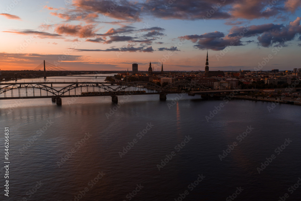 Fototapeta premium Riga, Latvia at sunset with St. Peter's Church spire, historic old town buildings, Vansu Bridge, and the Daugava River under a warm orange sky.