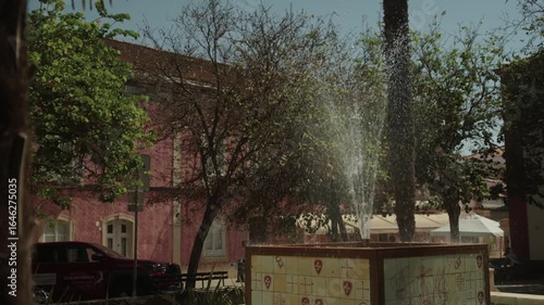 Water fountain spraying in a sunny square with pink building in the background, Portugal