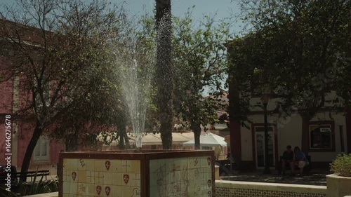 Water fountain spraying in a sunny Portuguese town square with tiled base and trees