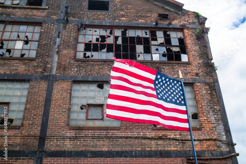 American flag waves in front of an abandoned factory with broken windows, symbolizing the decline of U.S. manufacturing and its shift overseas amid U.S.-China trade tensions