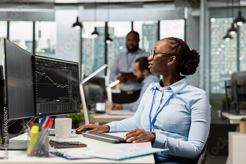 Trader in brokerage firm office analyzing market metrics and executing trades using trading app panel on PC. African american woman working in prop firm monitoring stock exchange