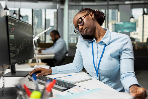 Brokerage company worker talking on phone while monitoring stock prices on computer screens. African american woman reviewing financial charts and making calls to execute trades