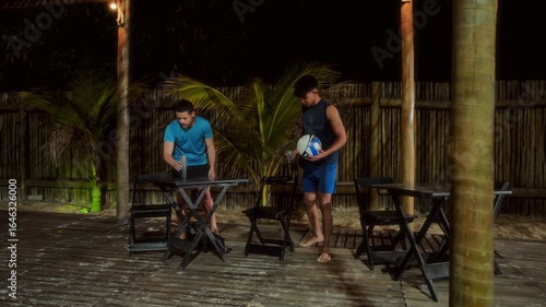 Two Men Relaxing and Laughing with Volleyball on Outdoor Wooden Deck at Night