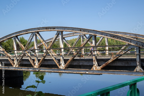 a weathered, rusty arched metal bridge spans a tranquil river on a sunny day. its intricate truss structure is reflected in the calm blue water, surrounded by green trees and a clear sky
