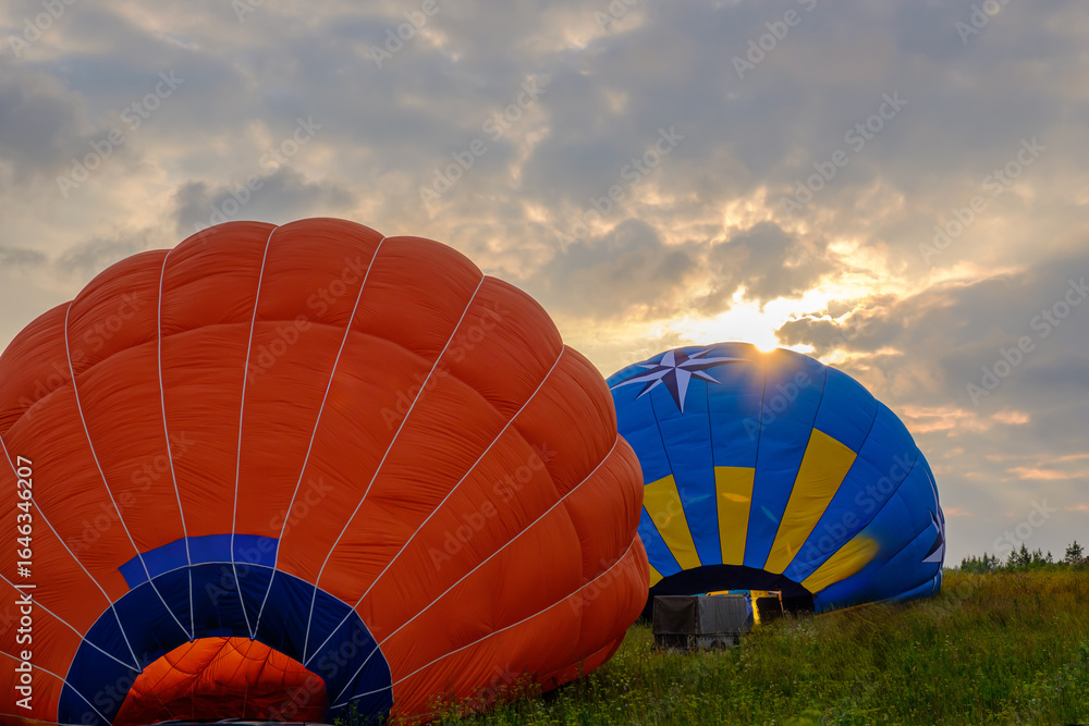 Obraz premium Colorful hot air balloons being inflated at sunrise in a scenic meadow, vibrant orange and blue fabric against dramatic cloudy sky, adventure travel and outdoor festival concept, panoramic photography