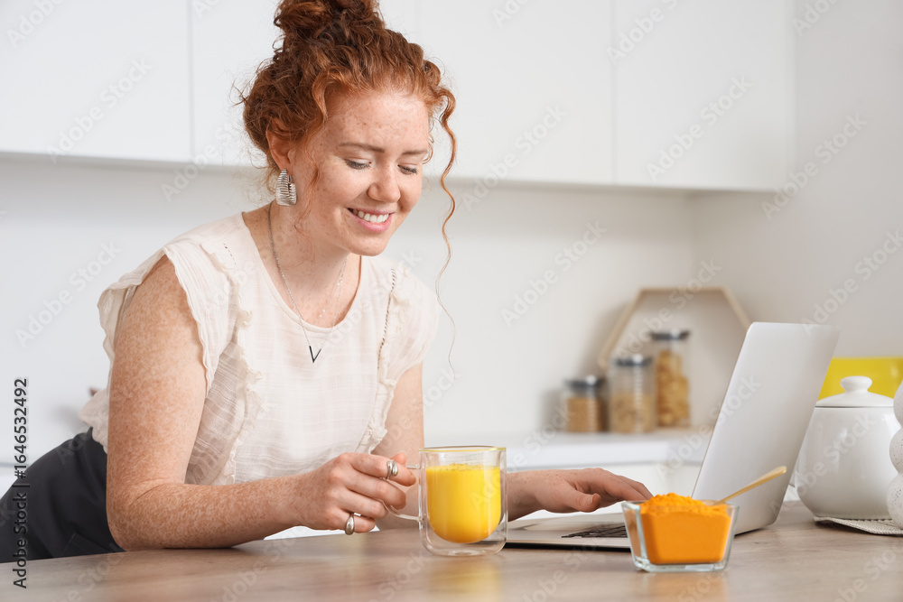 Young redhead woman with turmeric drink using laptop at table in kitchen Young redhead woman with turmeric drink using laptop at table in kitchen