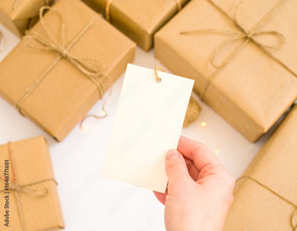 Fototapeta premium Hand Holding Blank Gift Tag Mockup: Wrapped Gifts Background. Gift-Giving, Celebrations & Thoughtful Branding