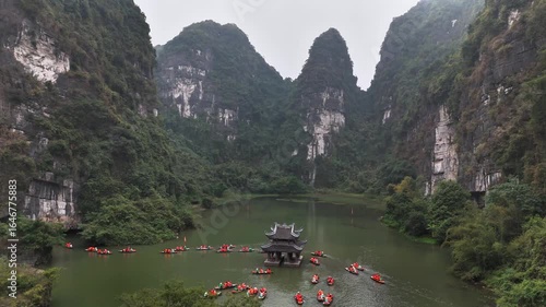 Sampan boats surround a traditional riverside pavilion set between towering limestone cliffs in the UNESCO-listed Trang An Landscape Complex, Ninh Binh, Vietnam, captured by aerial drone shot.