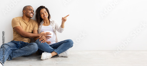 Smiling African American couple sitting on floor, man holding pregnant wifes belly while she points forward at copy space, imagining their future together, panorama