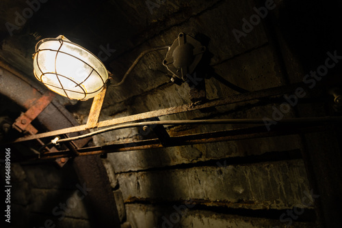 Quadro em tela Dim caged lamp illuminating a rusty steel frame and brick wall inside an abandon