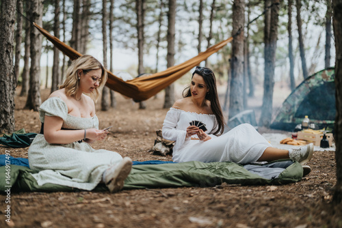 Two women sit comfortably in a tranquil forest setting, playing a card game near their camping site. The outdoor ambiance, with trees, hammocks, and camping gear, exudes relaxation and leisure time.