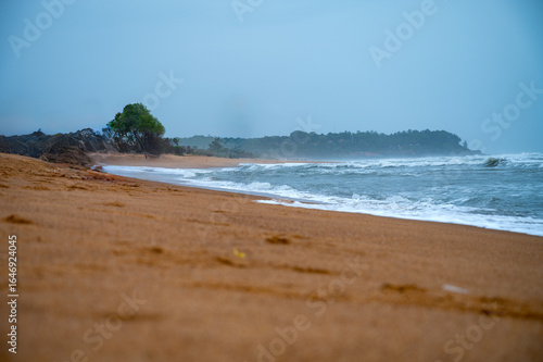 A tranquil coastal view of a pristine beach in Goa, India, captured during the monsoon season. Golden sand meets the foamy waves of the Arabian Sea under a soft blue sky