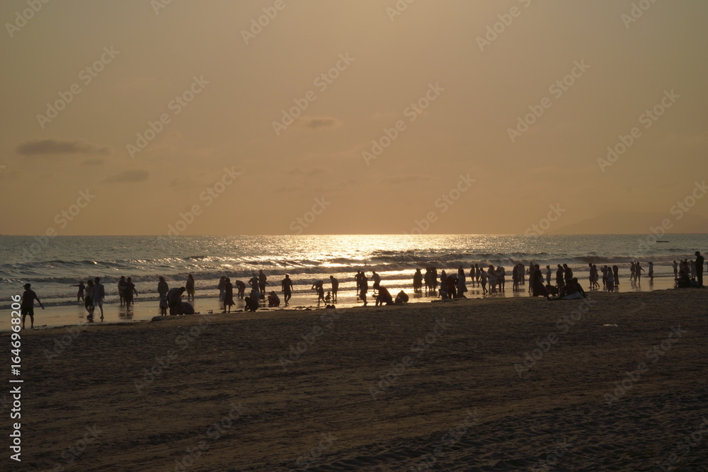 Obraz premium Golden Hour at a Crowded Tropical Beach - Vibrant Sunset Over Ocean with People Silhouettes and Scenic Coastal View
