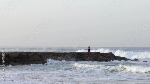 Wide shot of man standing on rocky jetty watching a surfer ride a wave. Cold clear morning, filmed in 4K at 50fps.