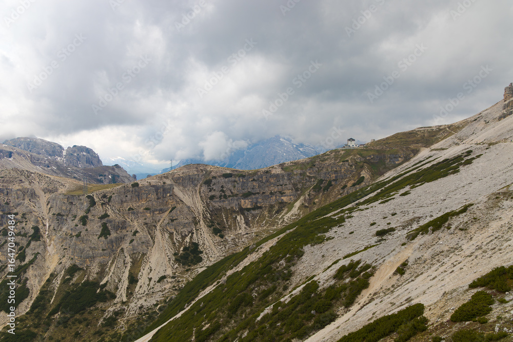 Fototapeta premium Panoramic view from Tre Cime di Lavaredo with distant mountain refuge, dramatic cliffs, cloudy sky, and rugged alpine slopes in the Dolomites.