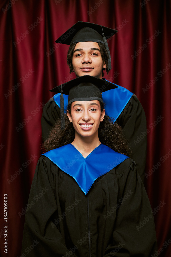 Obraz premium Portrait of young adult Black man and young adult biracial woman wearing graduation caps and gowns standing in photo booth, smiling at camera celebrating academic achievement together
