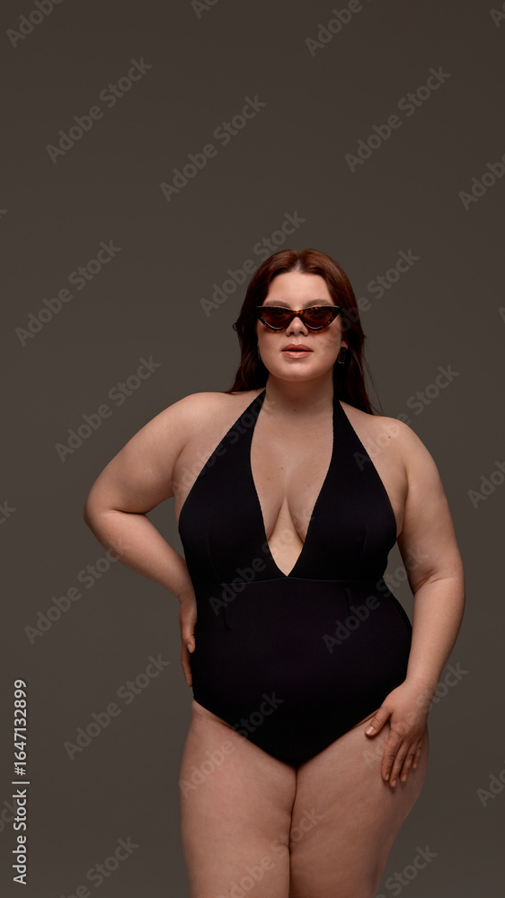 Obraz premium A plus-size woman confidently models stylish black swimwear paired with chic sunglasses. Concept of body positivity and summer fashion inclusivity.