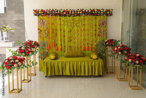 Mehndi ceremony backdrop with green drapes, floral strings, and decorative flower text, featuring elegant couch seating for traditional Indian wedding celebrations.