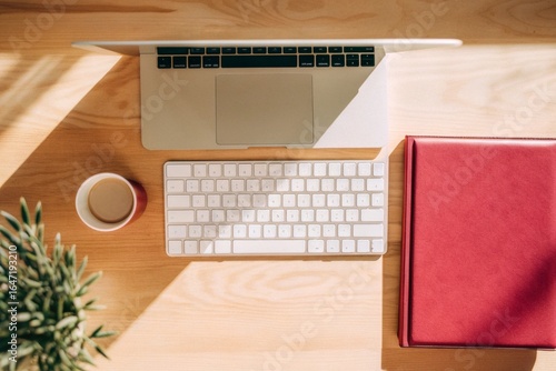 Home Office Desk Setup: Overhead view of a laptop, wireless keyboard, coffee mug, and a small plant on a light wooden desk