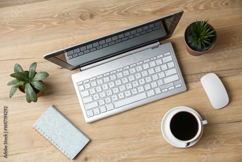Home Office Desk Setup: Overhead view of a laptop, wireless keyboard, coffee mug, and a small plant on a light wooden desk