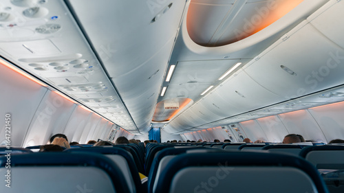 Interior view of a commercial airplane cabin with passengers seated during flight, showing rows of seats, overhead compartments, and soft ambient lighting