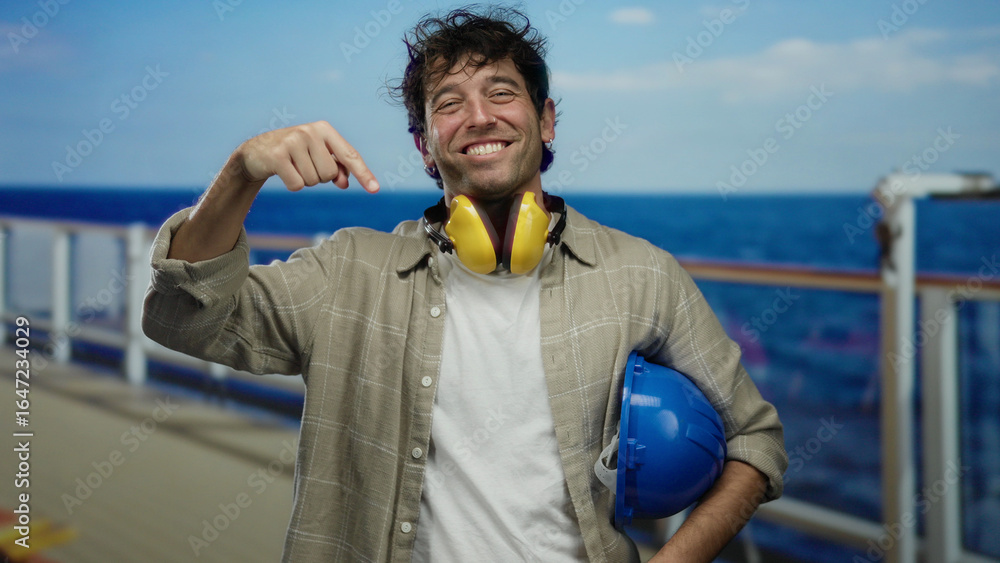 Fototapeta premium Hispanic man smiling on a cruise ship deck, wearing noise-canceling headphones and holding a blue hardhat with the ocean in the background.