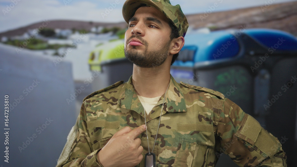 Fototapeta premium Young man points finger to dog tag on military uniform in street next to large recycling bins; pride duty.