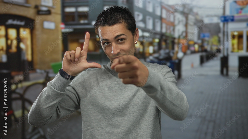 Obraz premium Young hispanic man making loser gesture with hand and pointing at camera in busy urban street; mockery.