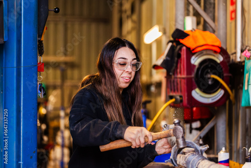 Female mechanic striking vehicle drive shaft with a hammer working on car repairs in workshop