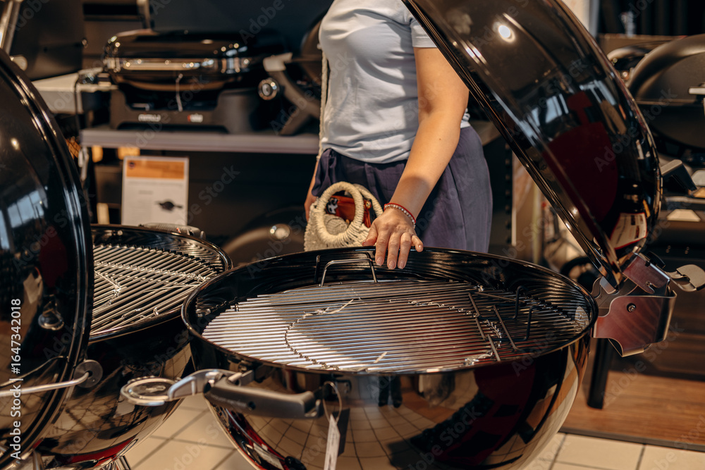 Obraz premium Woman inspecting modern charcoal grill in a retail store, showcasing sleek design and functionality for outdoor cooking enthusiasts